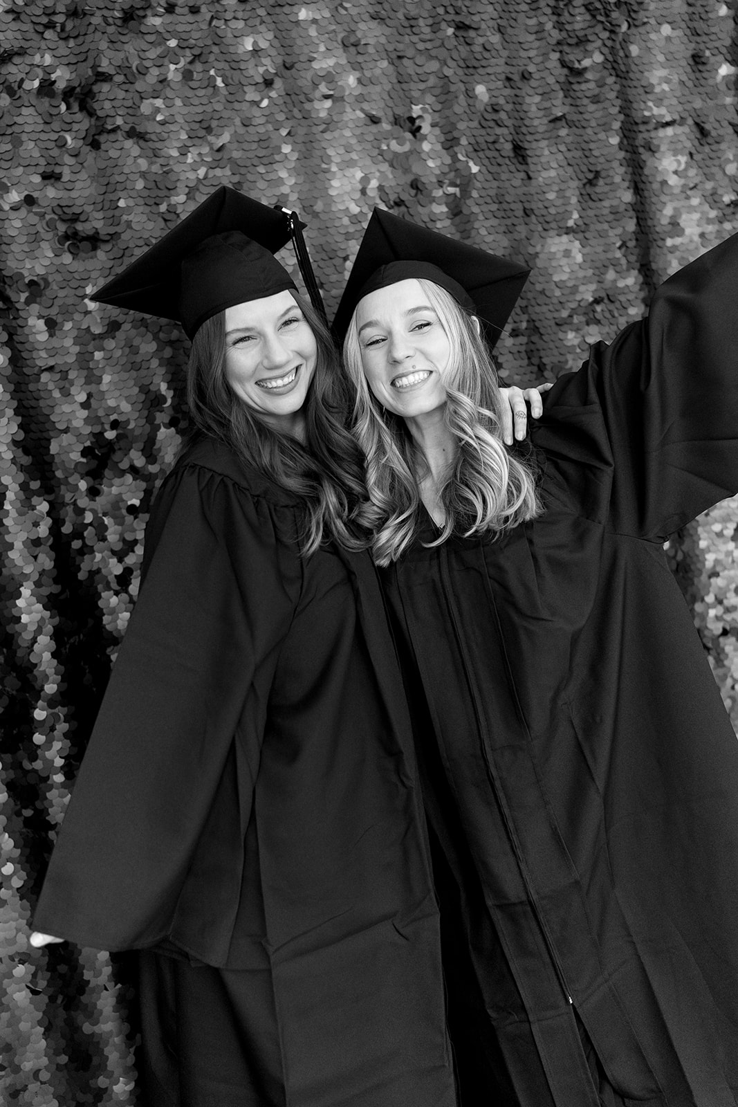 Two graduates in caps and gowns smile, one with an arm around the other, before a sequin backdrop, in black and white