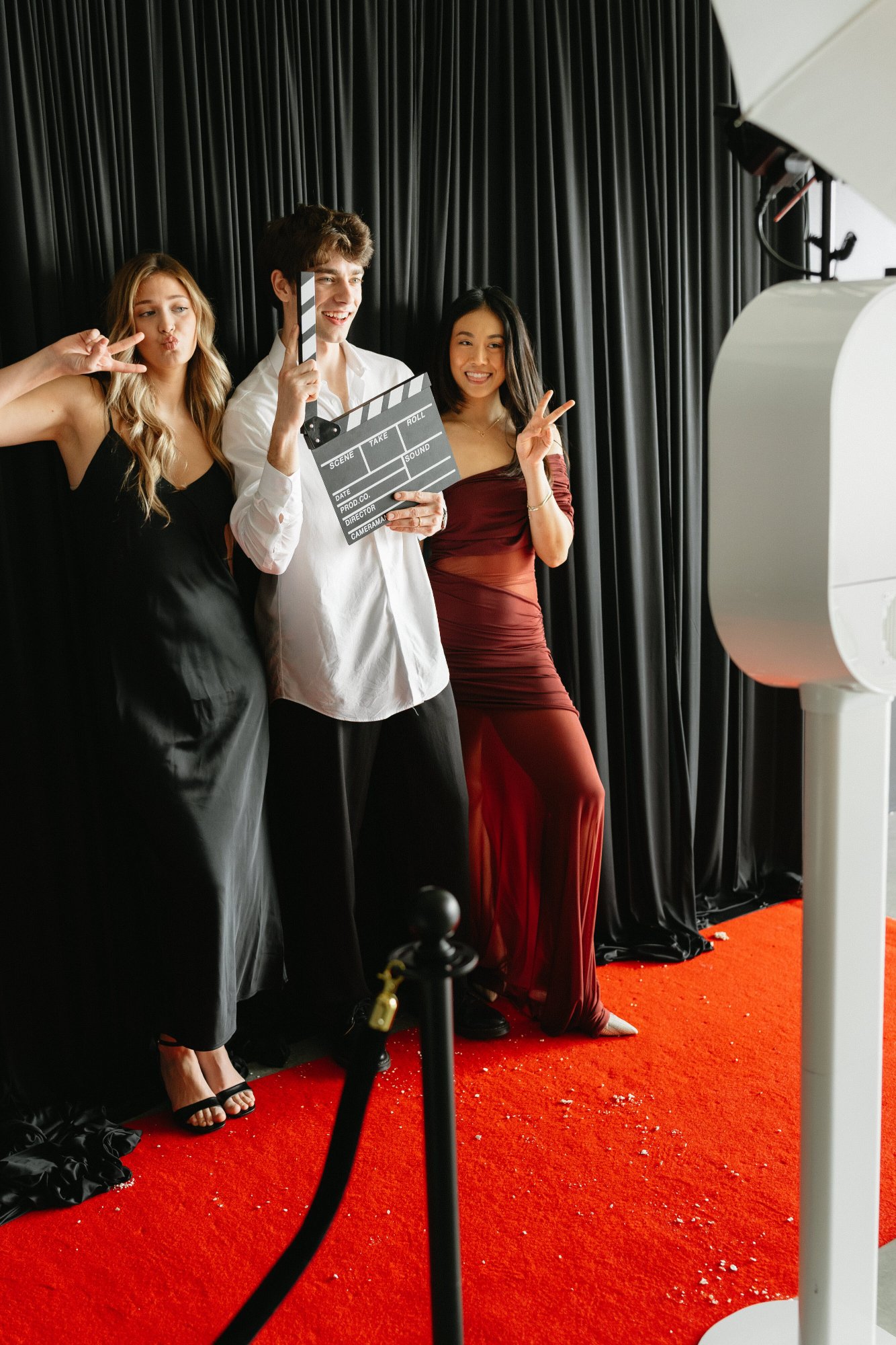 Three guests pose on a red carpet beside a white photo booth, one holding a clapperboard prop
