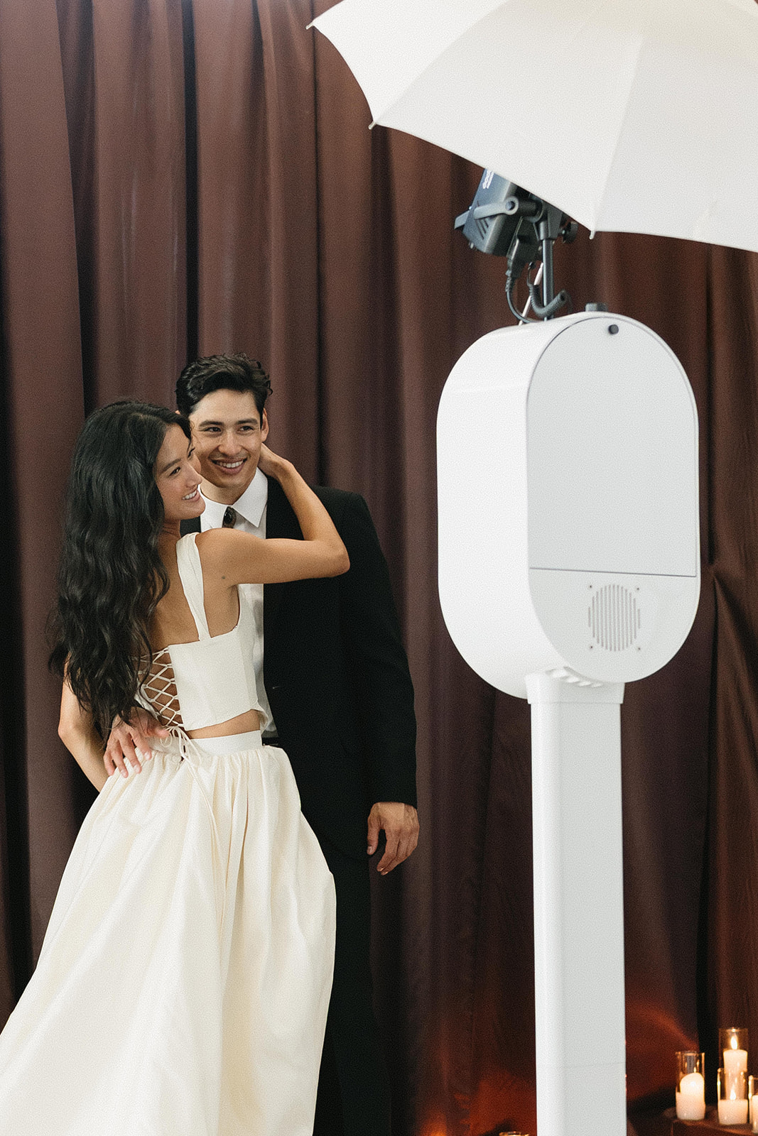 A couple in wedding attire posing at a white photo booth under an umbrella light, brown curtains behind